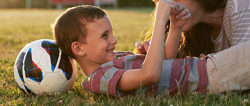 A boy with a soccer ball lying on a grassy field, smiling at his mother